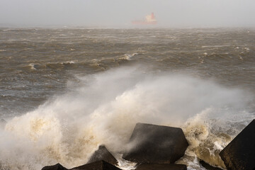 Orange ship sails out of harbor during a storm with rocks and waves in the foreground