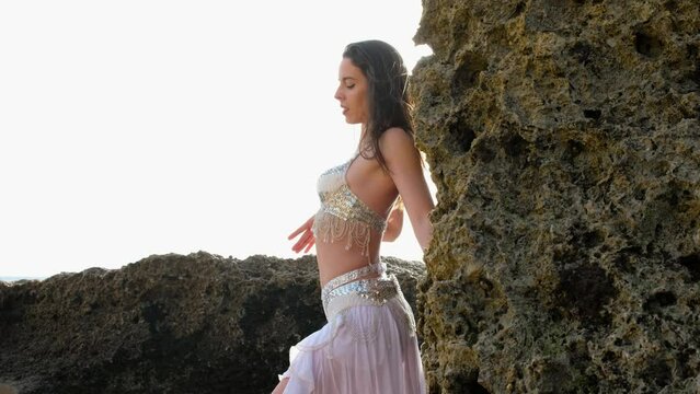 A beautiful woman performing a sensual belly dance in the sand on the beach. Spectacular landscape with blank holes and big rocks. Silhouette.