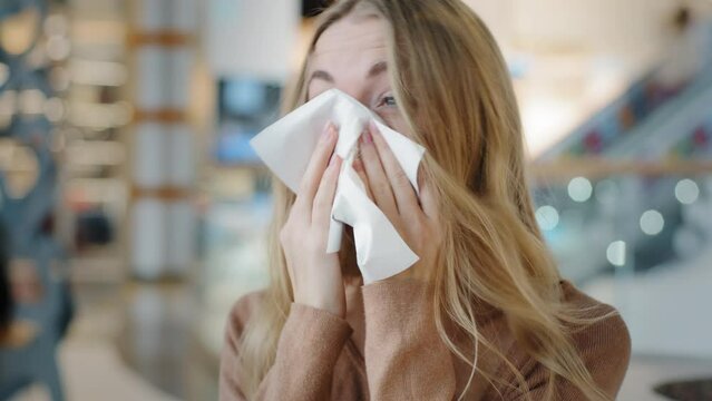 Portrait young caucasian woman with rhinitis runny nasal virus illness sneezes wipes with paper napkin in shopping mall student girl suffer from seasonal allergy dusty nose irritation disease symptoms