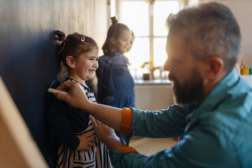 Teacher drawing little girl with chalks on blackboard wall indoors in playroom.