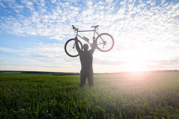 Fototapeta na wymiar A cyclist holds a bike raised up in his hands in a field at sunset.