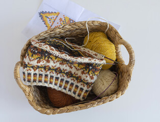 Fair Isle wool knitted hat in progress on the needles with yarn balls and knitting pattern in a wicker basket