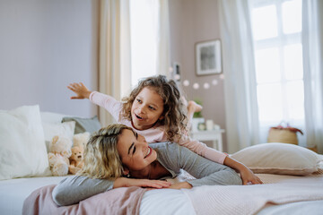 Obraz premium Happy little girl lying on mothers back on bed and looking at camera at home.