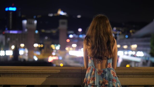 Tourist woman watch to city at night, blurred Avinguda Reina Maria Cristina perspective. Long haired lady stay in light dress, nice city lights seen in blur at background