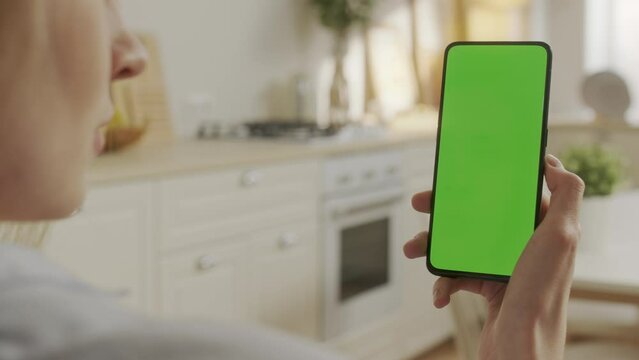Back View of Young Woman at Home Sitting on Kitchen Room With Green Mock-up Screen Smartphone. Female is Watcing Content Without Touching Gadget Screen. Modent Technology