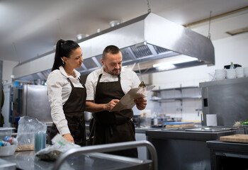 Chef and cook discussing menu indoors in restaurant kitchen. - obrazy, fototapety, plakaty