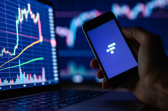 Vladimir, Russia - January 2022: FTX cryptocurrency trading application on a smartphone in hand against the background of monitors with financial charts. Selective focus on the logo.