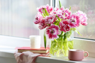 Vase with beautiful tulip flowers, cup of tea, book and candle on windowsill