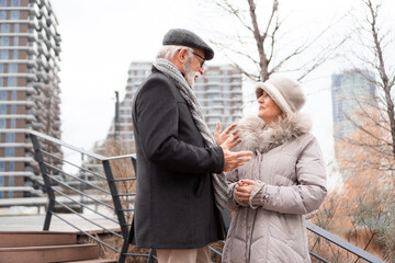 An elderly couple is walking around the town. The couple is smiling and looking happy in love.
