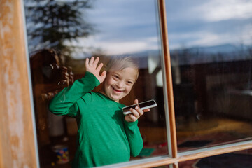 Happy little boy with Down syndrome using smartphone and waving through window