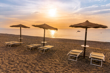  empty beach during beautiful sunrise or sunset with chaise loungues and nice umbrellas with blue sea, sun glow and amazing cloudy sky on thr background