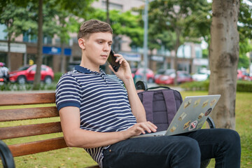 Young caucasian man sitting on the bench, working on the laptop and making