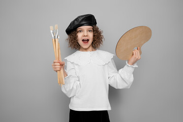 Emotional little girl artist poses in studio holds paintbrushes and palette. Children hobby concept