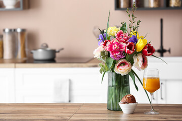 Vase with flowers, muffin and glass of juice on wooden kitchen counter