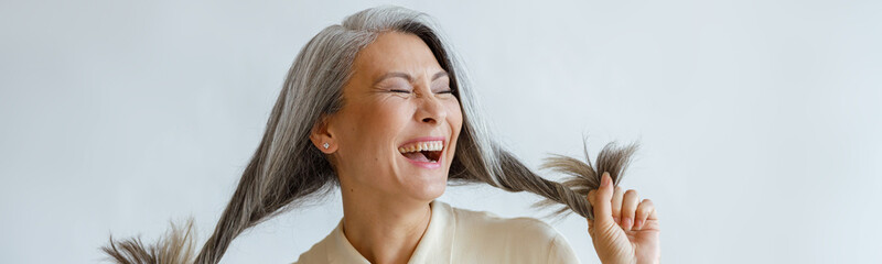 Joyful Asian lady in blouse holds long hoary hair laughing on light grey background in studio....