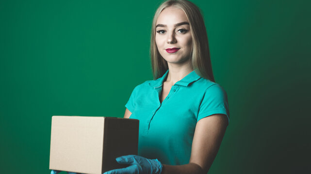 Beautiful Caucasian Female Standing Isolated On A Green Colored Background, Holding A Brown Parcel Box. Wearing Blue Surgical Gloves And Red Lipstick, Smiling Looking Straight At The Camera.
