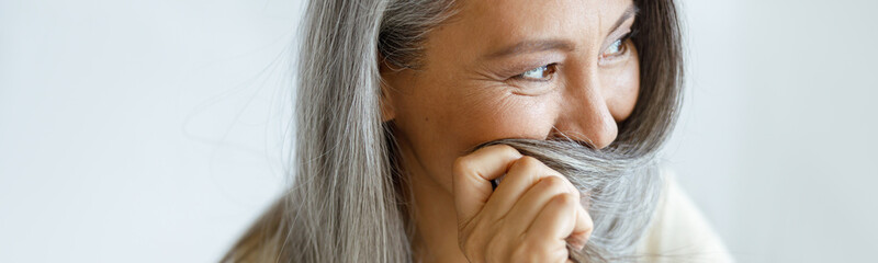 Playful middle aged Asian woman hides face with beautiful grey hair sitting on light background in studio. Mature beauty lifestyle