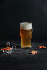 On a dark background, there is a filled glass goblet with a foamy alcoholic drink beer and shrimps lie on ice cubes