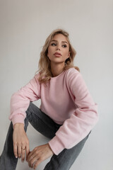 Fototapeta premium Beautiful young woman hipster with blond hair in a fashion pink sweatshirt and vintage gray jeans is sitting on the floor in the studio