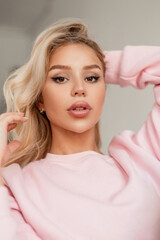 Young beautiful glamorous blonde woman with big sexy lips and brown eyes in a fashionable pink sweatshirt looks at the camera indoors