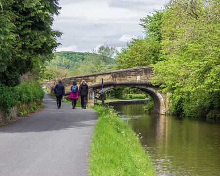 Three young women stroll along the tow path of the Leeds and Liverpool Canal at Dowley Gap near Bingley in West Yorkshire enjoying the peaceful waterway and admiring the stone bridge