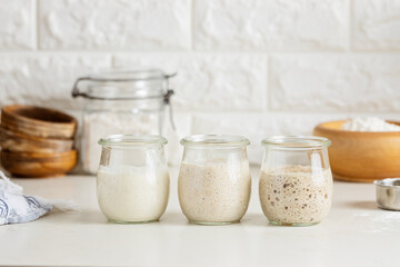 Three glass jars with active sourdough starter. Spelt, rye, and wheat. - obrazy, fototapety, plakaty