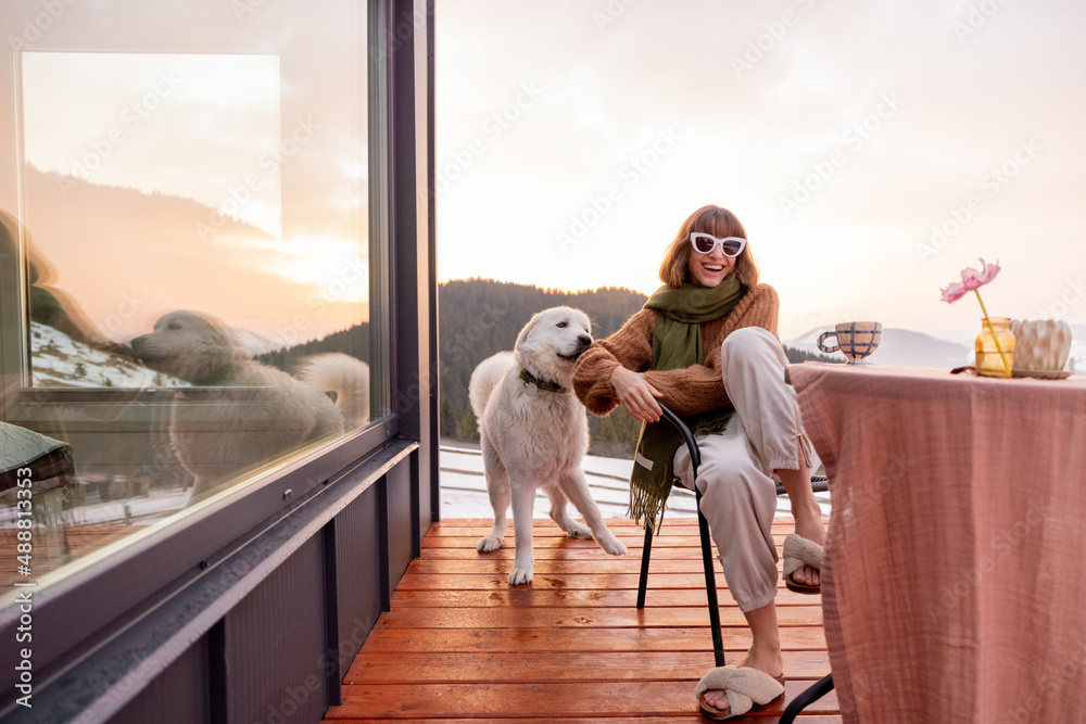 Wall mural Woman playing with her dog while resting on terrace of tiny house in the mountains, enjoying beautiful landscape during sunrise. Concept of small modern cabins for rest and escape to nature