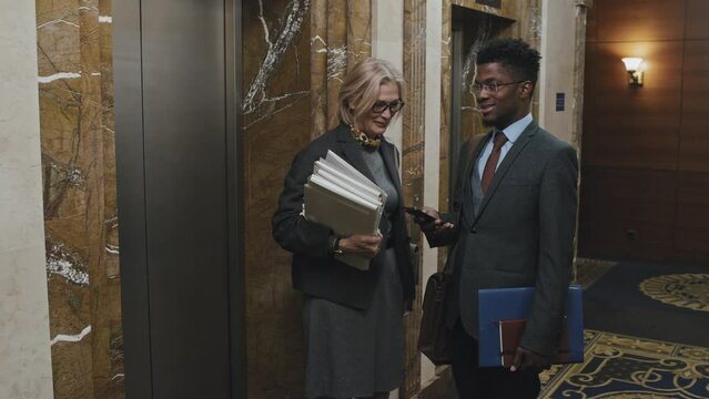 Young African American businessman and middle aged Caucasian businesswoman standing together by elevator in lobby of office center, speaking and greeting colleague who is passing by them