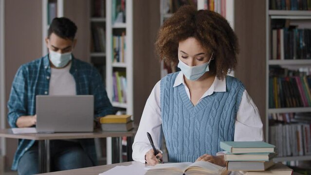 Young girl student in medical mask sits at desk in library preparing to lesson writing note in notebook looking at camera showing thumbs up sign approval guy studying on laptop students study in class