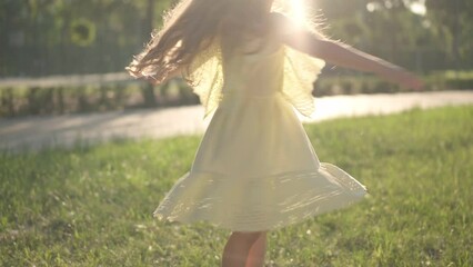 Unrecognizable little girl in yellow dress spinning in sunrays outdoors smiling. Excited happy Caucasian child having fun in spring summer park at sunset in sunbeam. Childhood and happiness concept - Powered by Adobe