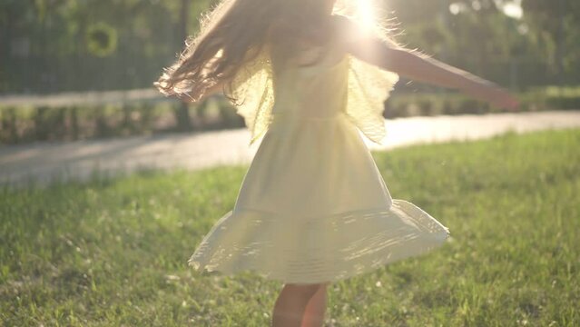 Unrecognizable little girl in yellow dress spinning in sunrays outdoors smiling. Excited happy Caucasian child having fun in spring summer park at sunset in sunbeam. Childhood and happiness concept