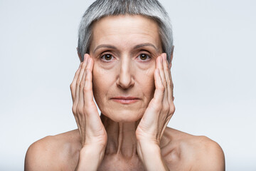 middle aged woman with grey hair touching face with wrinkles isolated on grey