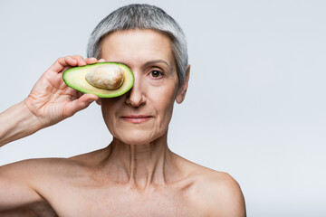 middle aged woman covering eye with half of avocado isolated on grey