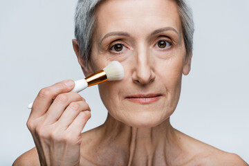 mature woman with bare shoulders applying face powder on cheek with cosmetic brush isolated on grey