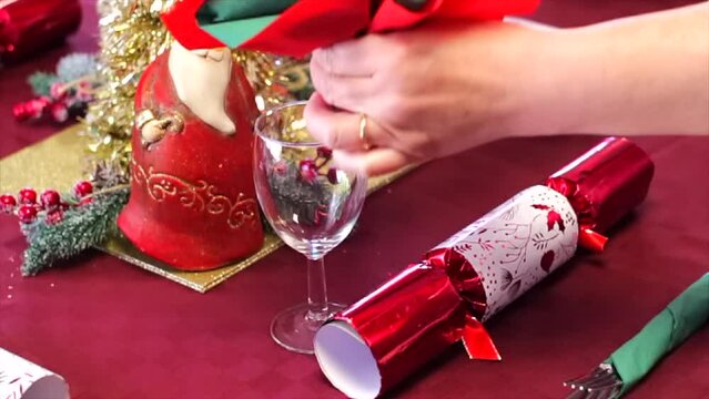 White womans hands filling wine glass with red and green napkins in a flower shape surrounded by Christmas ornaments and decorations. Slow motion origami. 