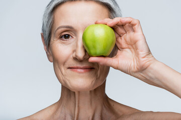smiling middle aged woman covering eye with green apple isolated on grey