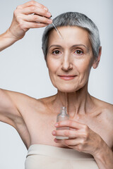 cheerful middle aged woman with grey hair holding bottle and applying serum isolated on grey