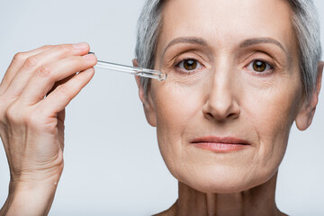 close up of middle aged woman with grey hair applying moisturizing serum isolated on grey