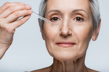 close up view of pleased middle aged woman with grey hair applying moisturizing serum isolated on grey