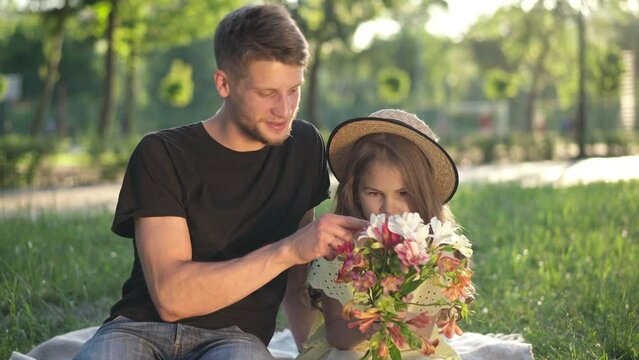 Little girl admiring bouquet of flowers sitting with young man outdoors in sunbeam. Portrait of pretty Caucasian daughter and happy handsome father resting together on spring summer weekend in park