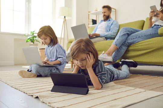 Modern family using different devices. Little child lying on floor at home and watching video on tablet while mum is using smartphone and brother and dad are using laptops. People, technology concept