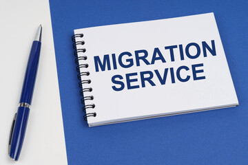 On a blue-white surface lies a pen and a notebook with the inscription - Migration Service