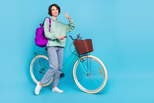 Photo of funky pretty woman wear green sweatshirt rucksack thumb up driving cycle empty space isolated blue color background