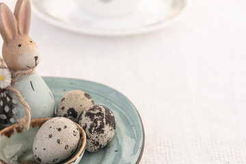Quail eggs and blue Easter rabbits on blue ceramic plate, white cup of tea or coffee on white table. Breakfast at Easter. Mock up