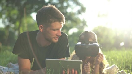 Caucasian young man using tablet lying with little girl wearing VR headset in sunbeam. Positive father enjoying weekend with daughter playing in augmented reality resting in sunny park outdoors