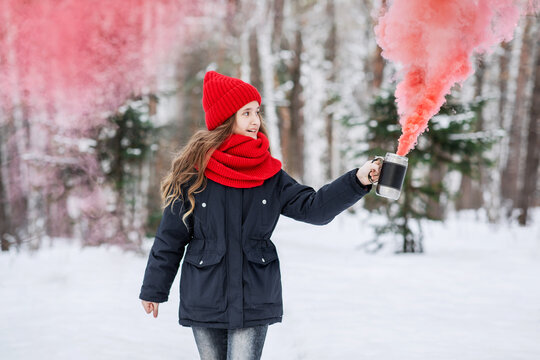 A teenage girl in red clothes plays with colored smoke on a walk in a winter park. Beautiful photos in a pine forest. Visual effects on the photo. Beautiful teenager in nature.