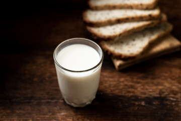 A full glass of milk. Bread in the background is blurred. Dark wooden background. Morning breakfast. Rustic style
