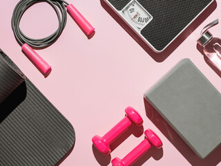 Stylish gray and pink home fitness flat lay. Top view of gray sport mat, yoga block, skipping rope, pink dumbbells, water bottle, scales on pink background. Set for pilates, fitness, yoga. Copy space