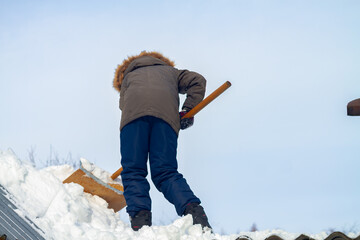 A teenage Caucasian boy cleans snow with a shovel from the roof of a house, sheds in winter or spring