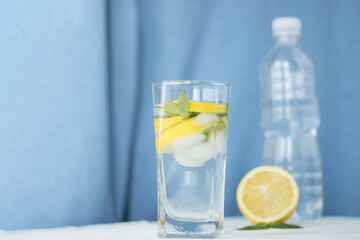 glass of water with lemon and ice and mint on blue background and white table